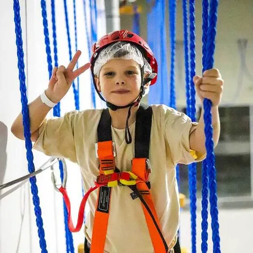 Мальчик в защитном снаряжении на канатах в развлекательном центре, демонстрирует знак мира, веселье