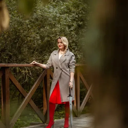 На мосту в парке женщина в красном платье и сером пальто с мечом в руках позирует на фоне зелёного ландшафта