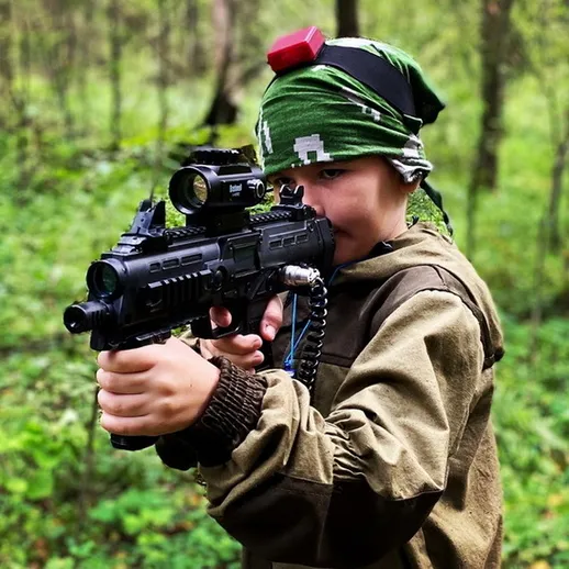 Мальчик в лесу с игрушечным автоматом, готовый к игре, одет в камуфляжную куртку и бандану, солнечный день