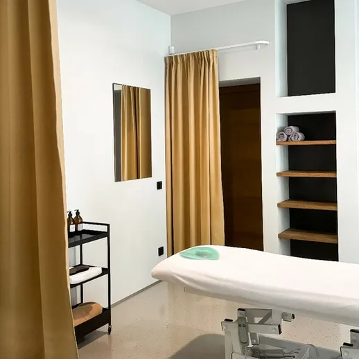 Уютный массажный кабинет с белоснежным массажным столом и серыми шторами в современном интерьере на фоне полок с полотенцами