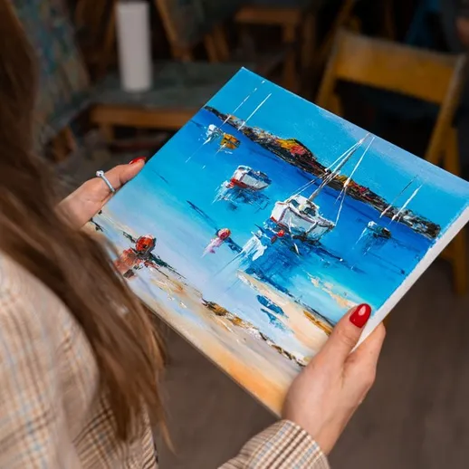 Художник держит яркую картину с морским пейзажем на фоне пляжа и лодок, написанную маслом в уютной студии