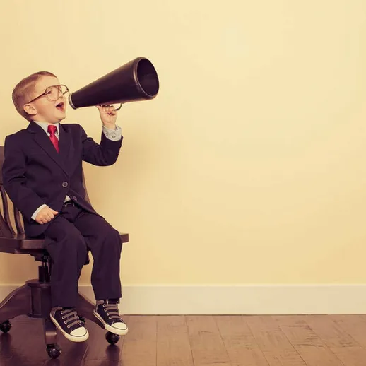 Мальчик в костюме с мегафоном сидит на стуле и радостно кричит в пустую стену