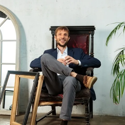 Молодой человек в костюме расслабленно сидит в кресле с перекрещенными ногами и улыбкой