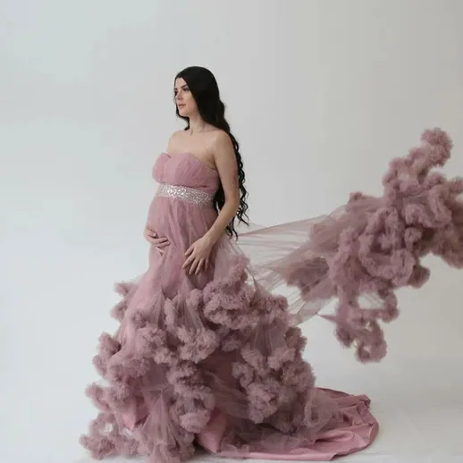 Беременная женщина в розовом платье позирует на нейтральном фоне с облаками ткани за спиной