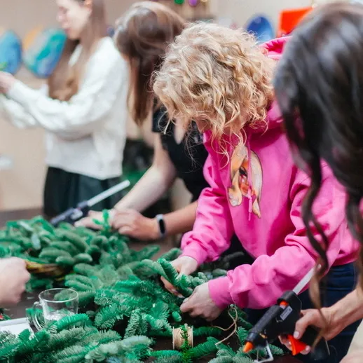 Женщины создают новогодние украшения в мастерской, используя искусственные еловые ветки и клей, в декабре