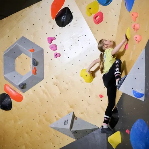 Юная скалолазка выполняет сложный маршрут на бoulder стенде в тренировочном зале на соревнованиях