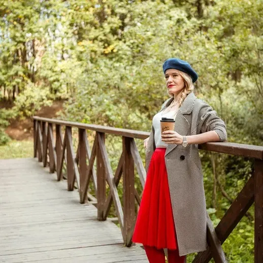 Женщина в сером пальто и красной юбке с кофе на мосту в лесу осенью