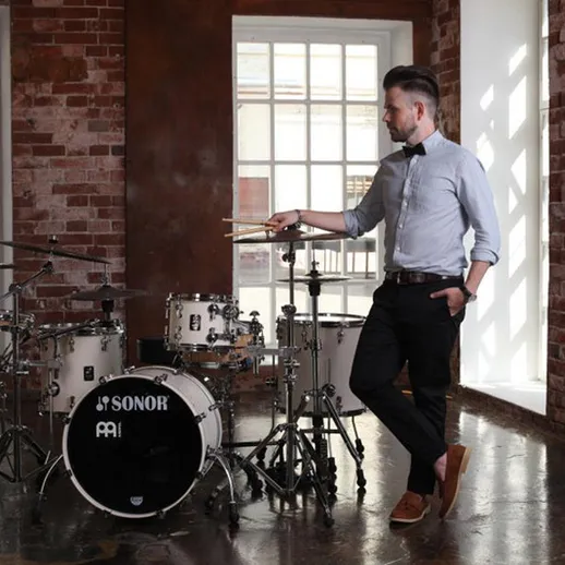 Молодой музыкант позирует рядом с барабанами в стильной студии с кирпичными стенами и большими окнами