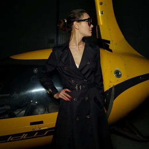 Женщина в черном пальто позирует рядом с желтым вертолетом в темном ангаре