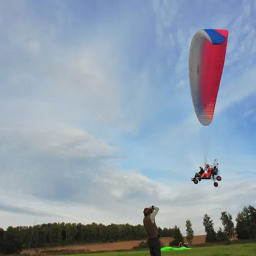 Парапланерист взлетает с поля в ясный день, в окружении зелени и облаков