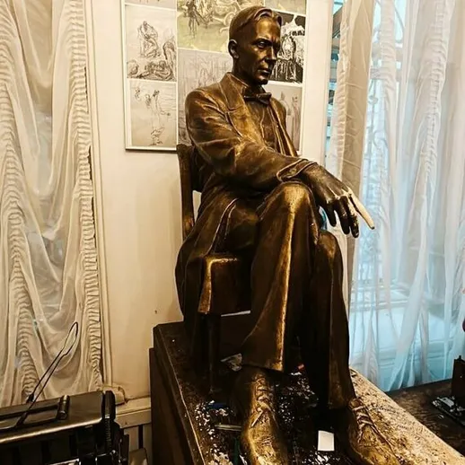 Статуя человека в костюме, сидящего на стуле в художественной галерее, окружённая эскизами и старинными предметами