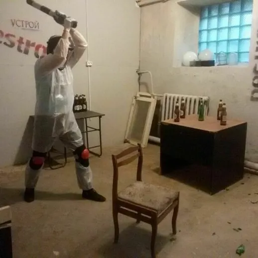 Мужчина в защитном костюме разрушает предметы в комнате с бутылками и мебелью