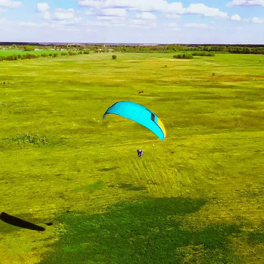 Парапланерист в синем параплане парит над зелеными полями в ясный день