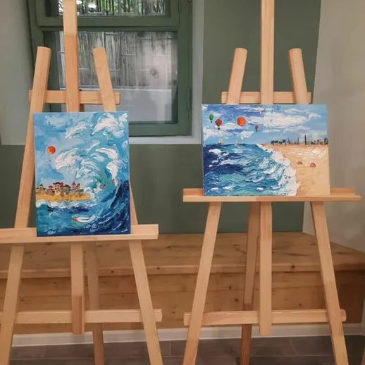 Две картины с изображением моря и пляжа, выставленные на мольбертах в уютном помещении