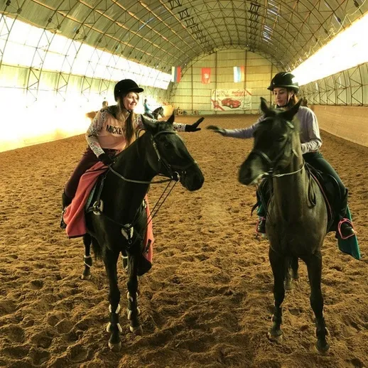 Две всадницы на черных лошадях обучаются верховой езде в манеже, освещенном солнечными лучами