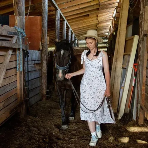 Девушка в светлом платье ведет черную лошадь по конюшне в солнечный день
