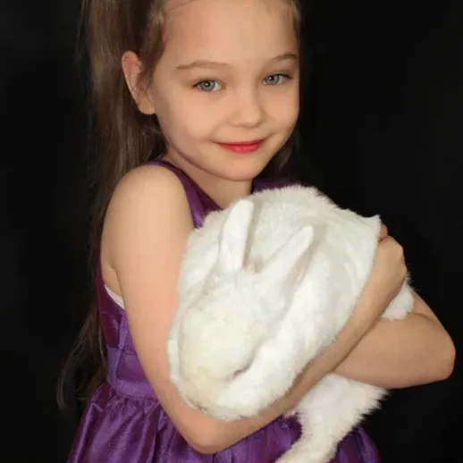 Девочка с белым кроликом на черном фоне, улыбается, одета в фиолетовое платье, вечерняя съемка