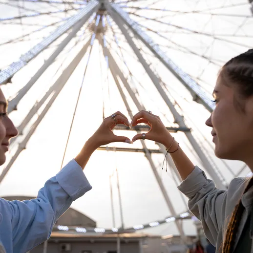 Две подруги делают сердце из рук на фоне колеса обозрения в парке аттракционов