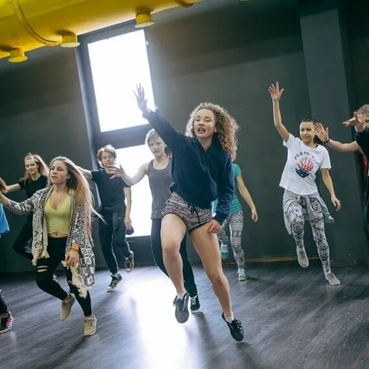 Группа танцоров в зале для репетиций исполняет энергичный танец под руководством инструктора, обсуждая движения и стили танца