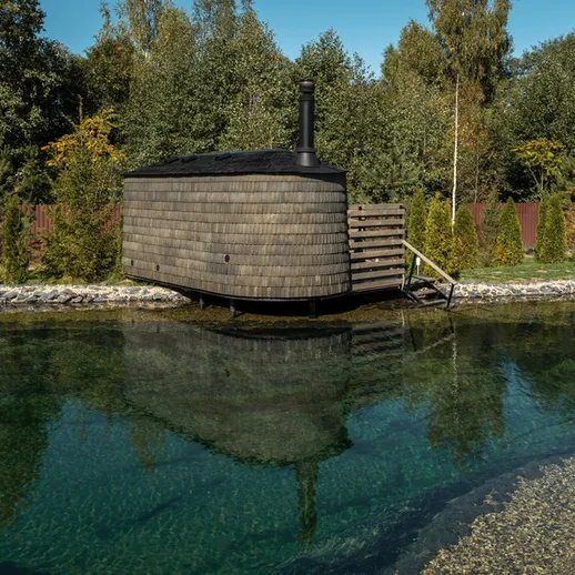 Сруб с сауной на берегу прозрачного водоема, окружённого зелеными деревьями в солнечный день