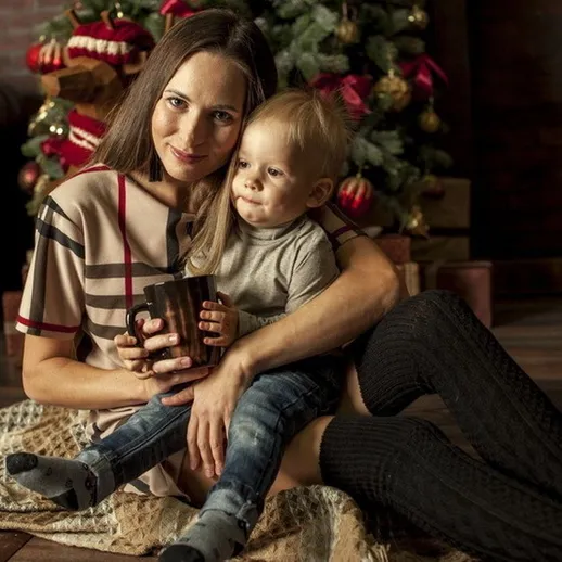 Мать с маленьким ребенком в уютной комнате у новогодней елки, наслаждающиеся горячим напитком в зимний вечер