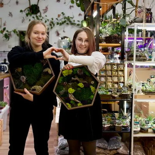 Две девушки в магазине растений демонстрируют экологичные террариумы с зеленью и искусственными фигурами в атмосфере уюта и гармонии