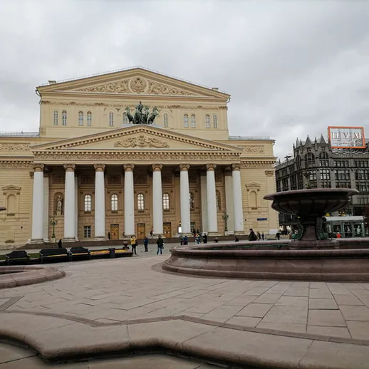 Здание Большого театра в Москве на фоне пасмурного неба, люди гуляют по площади и отдыхают на скамейках, вечер