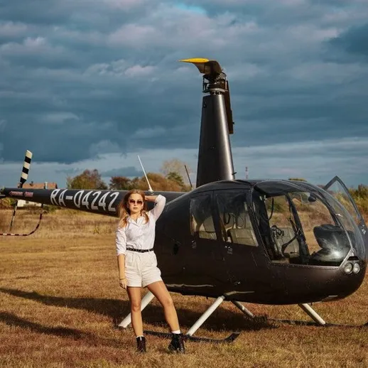 Девушка позирует рядом с вертолетом на открытом поле под серыми облаками