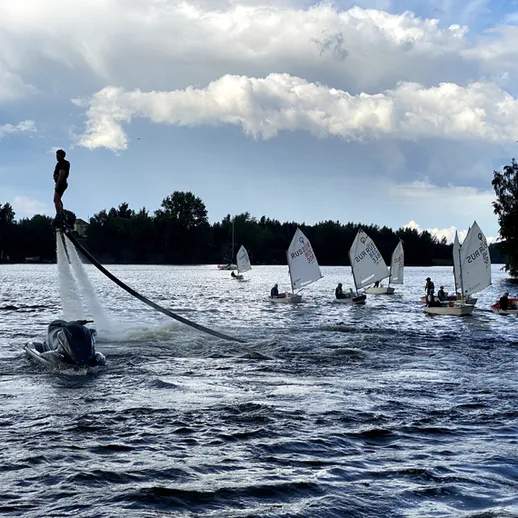 Человек на водном скейтборде выполняет трюки на озере среди парусных лодок в солнечный день