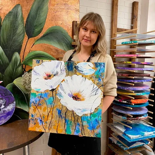 Художница держит картину с цветами в своей студии, на фоне других работ и зелёных растений