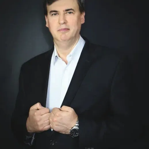 Строгий мужчина в темном костюме с часами на руке позирует на черном фоне в светлом свитере