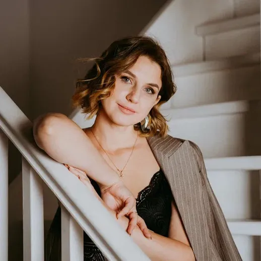 Женщина с вьющимися волосами позирует на лестнице в стильном наряде при мягком освещении в уютном интерьере