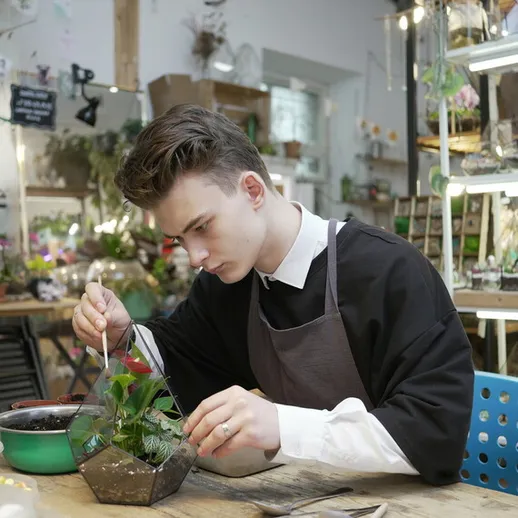 Молодой человек создает террариум в цветочном магазине, окруженный зелеными растениями и садоводческими инструментами