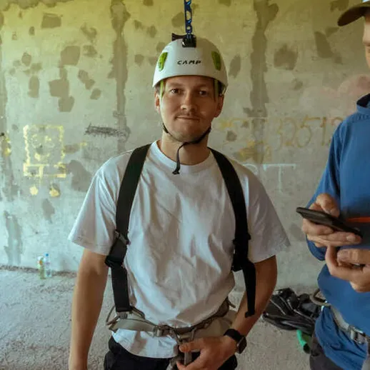 Молодой человек в каске и с альпинистским оборудованием готовится к спуску с высоты на строительном объекте