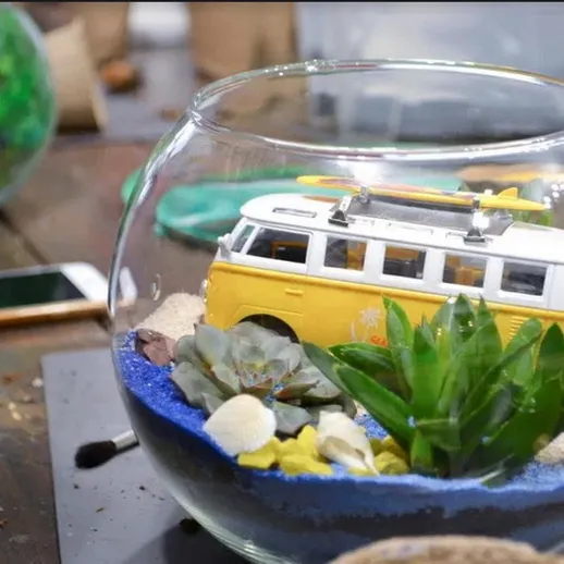 Красивый стеклянный контейнер с миниатюрным желтым фургоном и растениями на столе