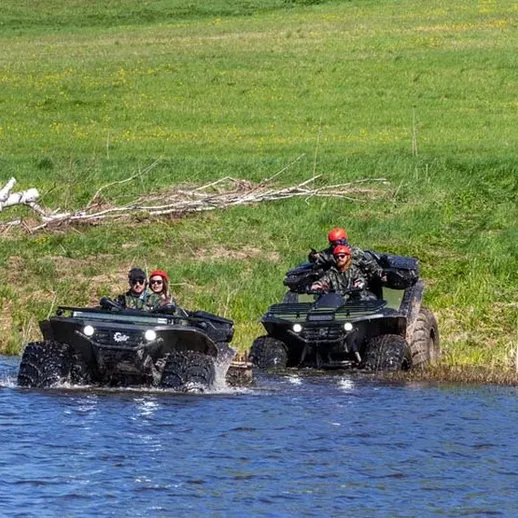 Два квадроцикла пересекают воду у берега реки на фоне зеленых лугов и деревьев в солнечный день