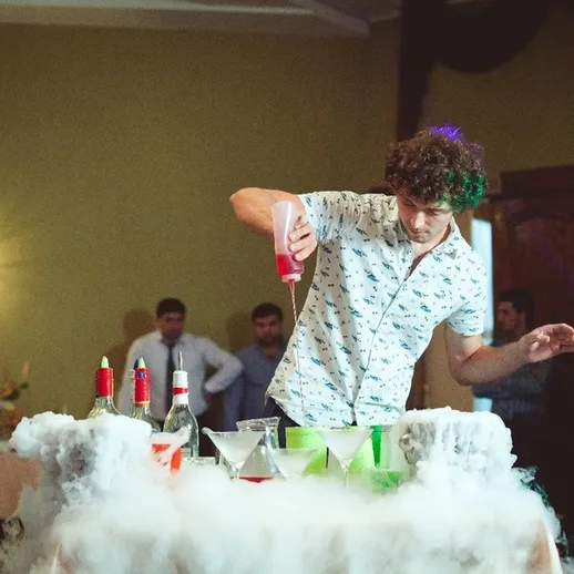 Молодой человек готовит коктейли на вечеринке в зале, окруженном дымом, с веселыми гостями вокруг