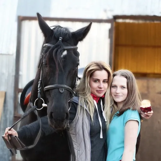 Женщина с ухоженной лошадью и девушкой на фоне конюшни в солнечный день